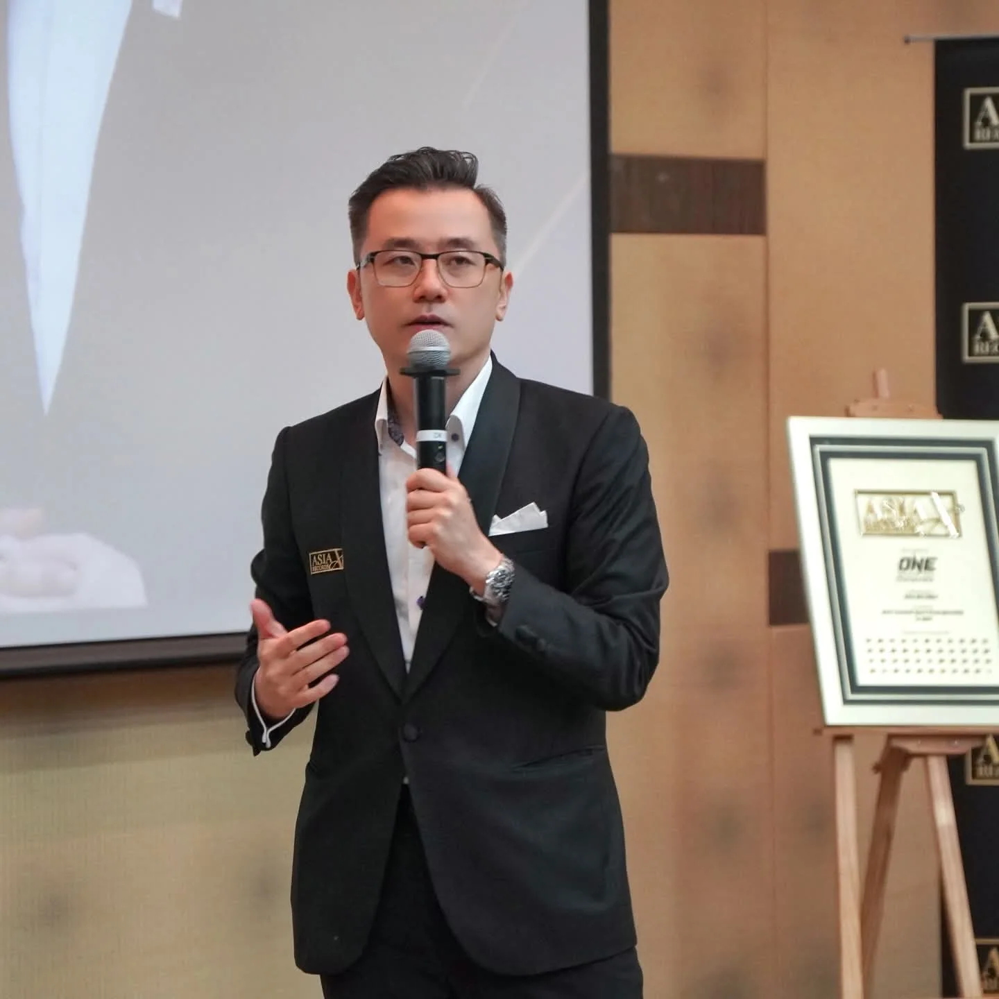 Dato' KK Chua speaking beside an achievement certificate